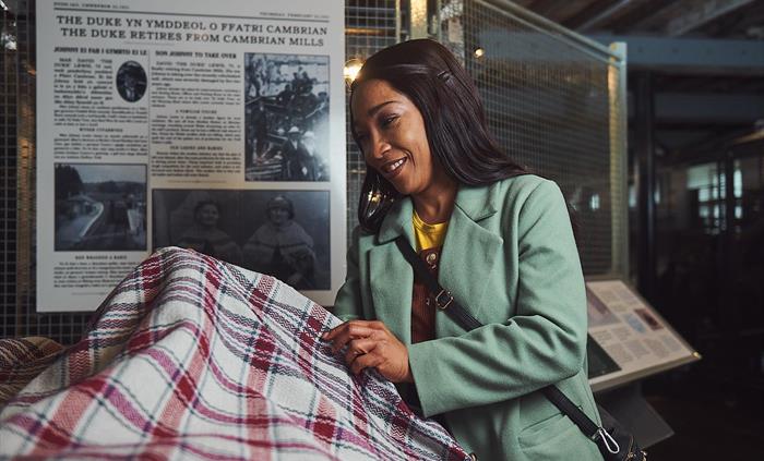 A lady in a green coat stands inside one of the weaving workshops. She is smiling and holding up a woollen blanket to examine it.
